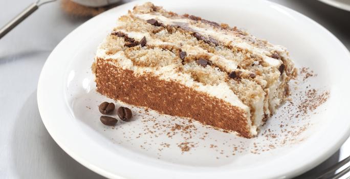 Fotografia em tons de branco, marrom e cinza, com prato branco ao centro com porção de bolo fatiado, no entorno garfo, peneirinha com chocolate em pó e pratos brancos empilhados, tudo sobre bancada em tons de cinza.