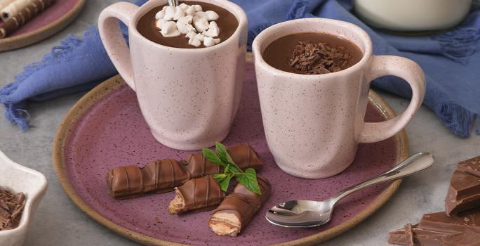Foto de duas canecas em tom rosa claro, contendo a receita de Chocolate Quente, sobre um prato em tom violeta, com um tecido em tom azul atrás, uma jarra com leite e pedaços de chocolate e canelas em pau sobre a mesa