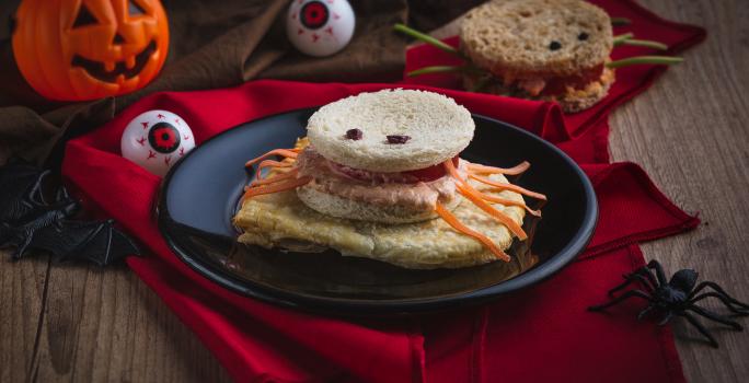 Imagem aproximada da receita de um sanduíche em formato de aranha, com pão, patas feitas com tiras de cenoura, além de o cenário estar decorado com itens de Halloween