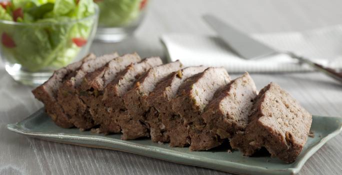 Fotografia em tons de marrom e cinza, com bolo de carne assada sobre peça de porcelana cinza e no entorno guardanapo branco com espátula por cima e dois potinhos com folhas verdes, tudo sobre bancada em tons de cinza.