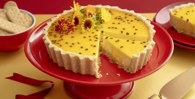 Foto da receita de Cheesecake de Maracujá e Chocolate Branco. Observa-se uma torta com calda de maracujá e flores comestíveis com um pedaço cortado, em cima de uma boleira vermelha.