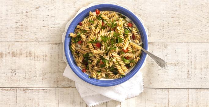 em uma mesa de madeira na cor cinza, contém um recipiente na cor azul que comporta a salada de macarrão.