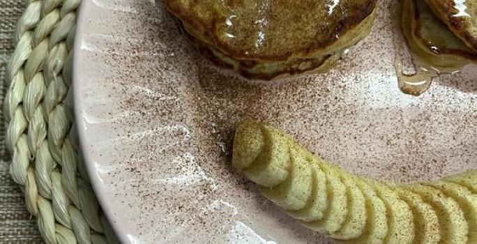 Foto da receita de Panquequinha de Aveia. Observa-se um prato com panquequinhas com mel e canela e do lado de baixo do prato, uma banana em rodelas para formar um sorriso.