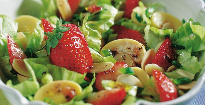 Fotografia em tons de verde em uma bancada de madeira azul com um recipiente redondo e fundo com a salada primavera composta por alface, cogumelo e morango.