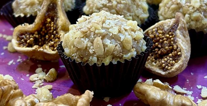 Foto de receita de Brigadeiro de Figo com Nozes. Observa-se o brigadeiro com nozes picadas em volta em uma forminha preta, com figos secos cortados ao lado e nozes na projeção da frente. Ao fundo, mais brigadeiros.