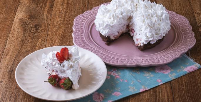 Imagem da receita de Torta Brownie, decorada com chantilly e morangos, servida em um prato rosa e ao lado há um prato menor branco com uma fatia. Tudo está sobre um pano decorado com motivos florais, sobre uma bancada de madeira