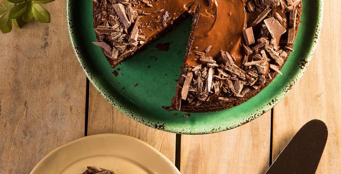 Fotografia em tons de verde em uma bancada de madeira clara com um suporte verde com o bolo de chocolate em cima. Ao lado, um prato com uma fatia do bolo e uma espátula para bolo.