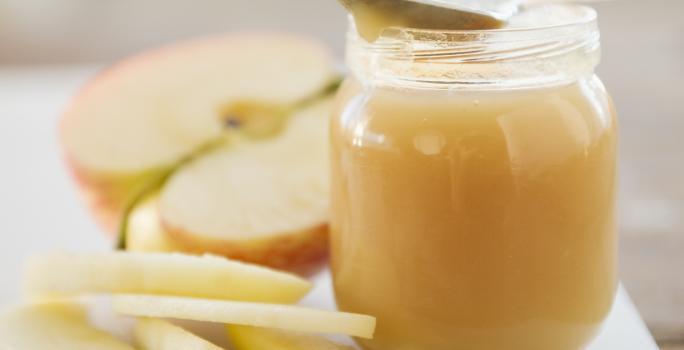 Fotografia de um potinho de vidro com um creme de maçã com iogurte e farofa doce. Apoiada no potinho, uma colher de sobremesa, e ao lado, cinco fatias de maçã sem casca junto com uma metade da maçã cortada ao meio, sobre uma tábua de cor branca.