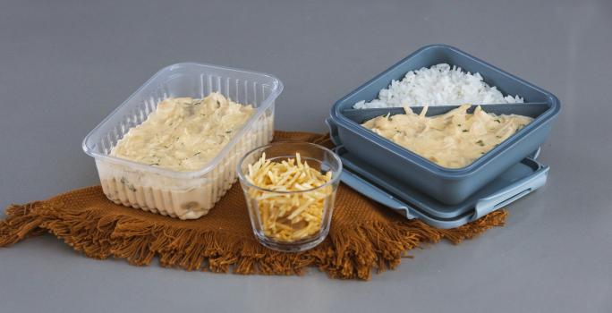 Foto da receita de fricassê de frango, servida em potinhos de marmita, com arroz e batata palha, sobre uma tábua de madeira em uma bancada cinza.
