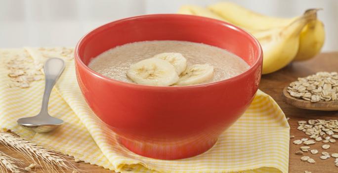 Fotografia em tons de amarelo e vermelho de uma bancada de madeira com um paninho amarelo e um recipiente redondo vermelho, sobre ele o mingau de farinha láctea com fatias de banana. Ao fundo duas bananas e aveias espalhadas pela bancada.