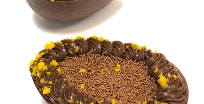 Foto em tons de branco, marrom claro, escuro e laranja. Na foto é possível ver uma bancada branca com duas cascas de ovos recheadas com brigadeiro, chocolate granulado e migalhas de bolo de cenoura