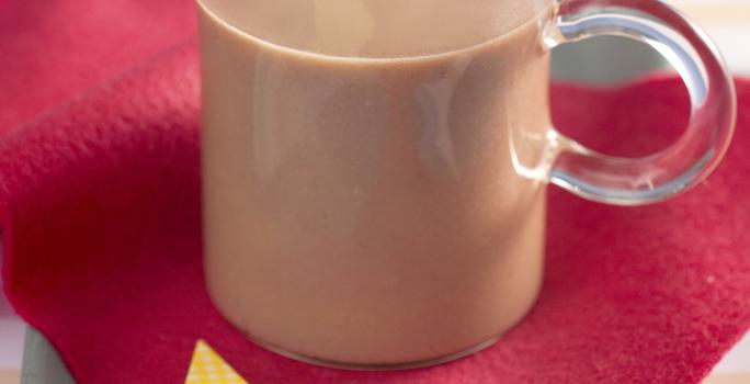 Fotografia em tons de vermelho e azul em uma bancada de madeira azul, uma toalhinha listrada em azul e rosa, um paninho vermelho com uma estrela amarela. Ao centro, uma xícara de vidro com chocolate quente.