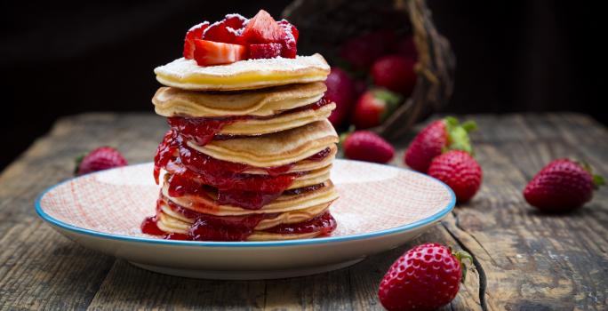 Fotografia de várias minipanquecas empilhadas com recheio de geleia de morango, e por cima, açúcar de confeiteiro e alguns morangos. As panquecas estão dentro de um prato fundo colorido, e ao fundo, morangos inteiros espalhados pela mesa de madeira.