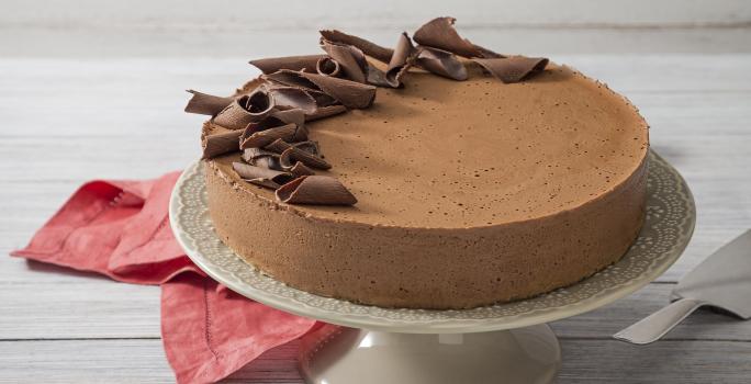 Fotografia em tons de branco e vermelho de uma bancada de madeira branca com um paninho vermelho, sobre ele uma bailarina branca com a torta mousse de chocolate decorada com raspas de chocolate.