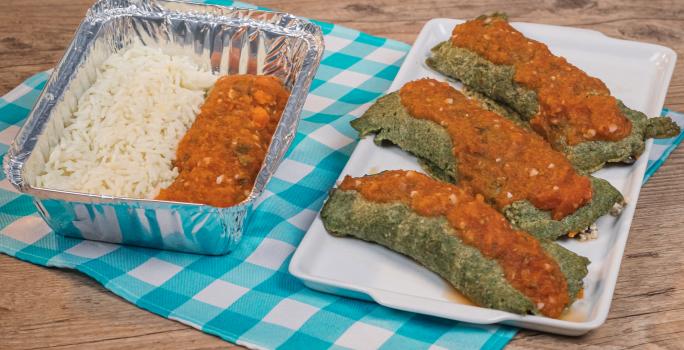 Foto de uma marmita de alumínio com arroz e panqueca verde dentro. Ao lado há um prato branco retangular com três panquecas verdes cobertas com molho de tomate. Tudo está sobre um pano azul e branco quadriculado numa mesa de madeira