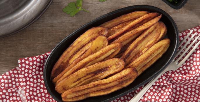 Fotografia em tons de vermelho em uma bancada de madeira, uma toalha vermelha com flores, um recipiente oval preto com várias bananas fritas dentro dele.