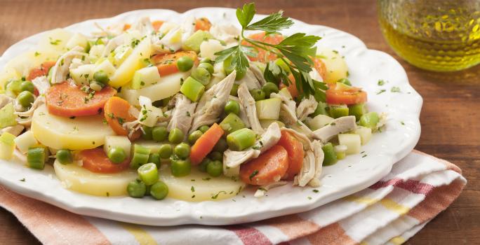 Fotografia em tons de marrom e branco de uma bancada de madeira com um paninho branco com listras vermelhas e amarelas sobre ele um prato branco redondo com bordas irregulares e a salada de frango com legumes. Ao lado um recipiente de vidro com azeite.