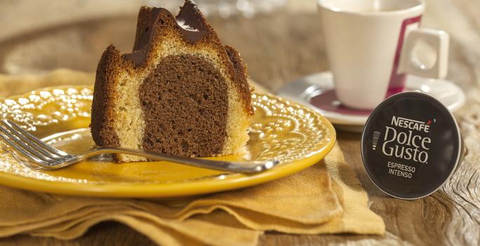 Fotografia em tons de marrom e amarelo de uma bancada de madeira com um paninho amarelo, sobre ele um prato redondo amarelo com uma fatia do bolo e um garfo. Ao fundo uma xícara branca com detalhes vermelhos e uma capsula de Dolce Gusto.