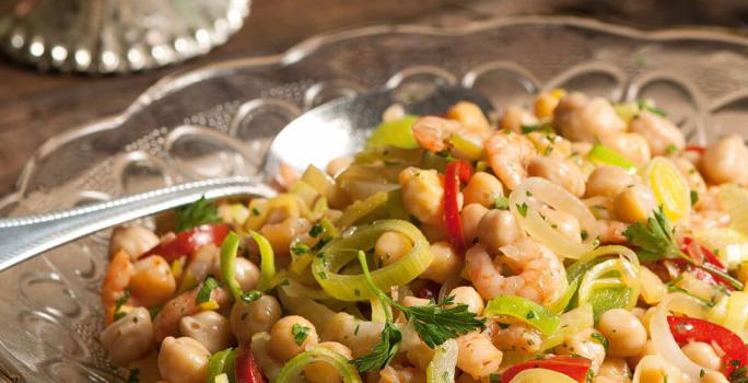 Fotografia em tons de bege e marrom, com salada de grã0-de-bico e legumes de cores verde e vermelha, dentro de saladeira de vidro com uma colher de servir, tudo sobre bancada de madeira.