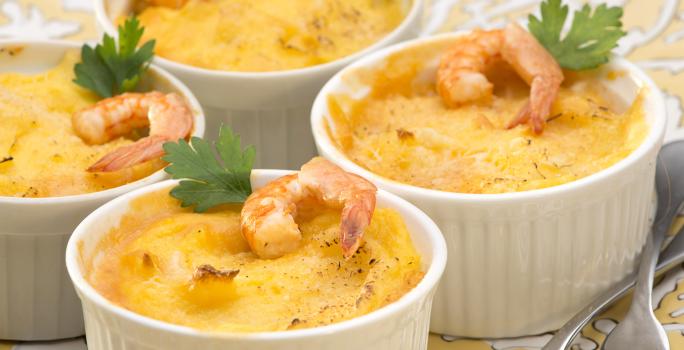 Fotografia em tons de branco e amarelo de uma bancada de madeira marrom, um paninho cinza e um prato amarelo com branco, sobre ele quatro recipientes redondo com o escondidinho de camarão decorados com um camarão e uma folha de salsinha.