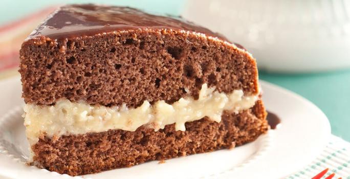 Fotografia em tons de branco, marrom e azul. Um prato branco contém uma fatia de bolo de chocolate com recheio de coco.