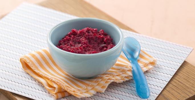 Fotografia em tons de roxo em uma bancada de madeira de cor marrom. Ao centro, uma tábua de madeira contendo um pano azul e um pano laranja, em cima há um recipiente contendo o purê. Ao lado, há uma colher azul.