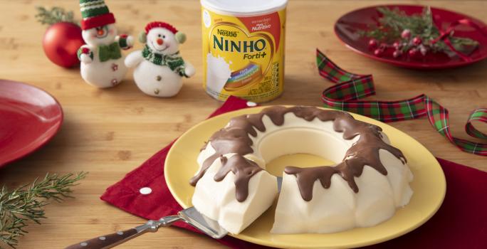 Fotografia em tons de vermelho e branco de uma bancada de madeira, um paninho vermelho e um prato amarelo com o bolo de sorvete com calda de chocolate em cima. Ao fundo decoração de natal, com mini bonecos de neve e uma lata de NINHO.