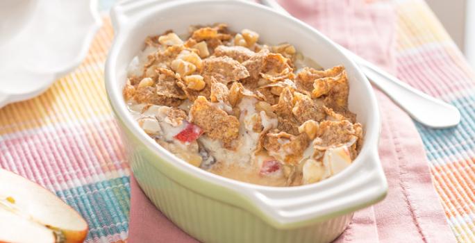 Fotografia em tons de rosa, pardo e verde claro de uma bancada de madeira parda vista de cima, um pano colorido nas cores rosa, azul e laranja. Um pano rosa e por cima um recipiente verde claro com alças que contém maçã, banana e cereal.