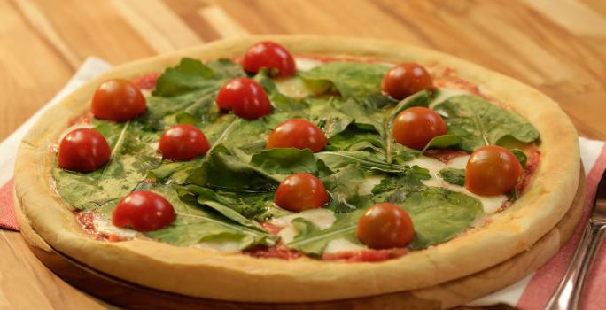 Fotografia em tons de dourado, verde e vermelho, ao centro pizza com folhas verdes e tomate-cereja sobre tábua redonda e guardanapo branco e vermelho, com talher ao lado, tudo sobre bancada de madeira em tons marrom.