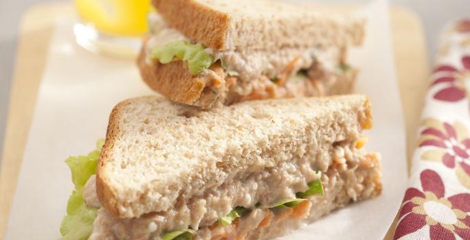 Foto em tons claros da receita de sanduíche de atum com queijo cottage servida em duas metades, uma por cima da outra, sobre uma tábua de madeira coberta com papel branco