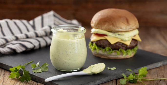 Fotografia em tons de verde em uma bancada de madeira clara, um pano branco com listras pretas, uma tábua de ferro preta e em cima dela, um potinho de vidro com a maionese verde caseira e ao lado um hambúrguer para acompanhar essa deliciosa maionese.