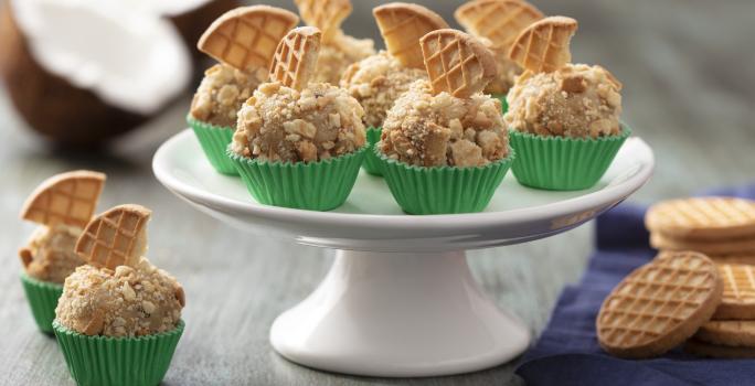 Fotografia em tons de cinza e verde de uma bancada cinza, com um prato branco alto com sete brigadeiros em forminhas verdes com pedaço de biscoito em cima. Ao lado dois brigadeiros, pano azul e bolachas.