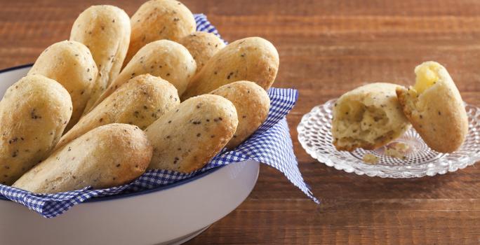 Fotografia em tons de dourado, branco e marrom, com recipiente branco contendo onze pãezinhos assados sobre guardanapo quadriculado em branco e azul, ao lado pratinho com pão aberto ao meio, tudo sobre bancada de madeira.