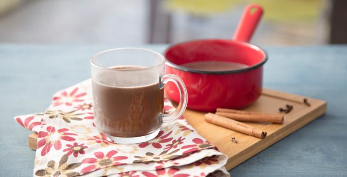 Fotografia em tons de vermelho e azul em uma bancada de madeira azul, uma tábua de madeira, um pano branco com flores rosas e marrons, uma panelinha vermelha pequena com o chocolate quente dentro dele. Ao centro, uma xícara de vidro com o chocolate quente