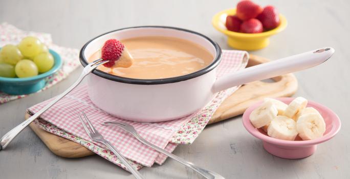 Fotografia em tons de branco, caramelo e rosa, tendo ao centro panela em ágata contendo doce de leite cremoso, garfinhos para fondue ao lado sobre guardanapo quadriculado e peça de madeira, no entorno potinhos com frutas picadas, tudo sobre bancada cinza.