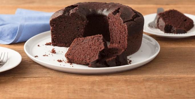 Fotografia em tons de marrom e branco de uma bancada de madeira clara com um paninho azul claro e um prato branco redondo com o bolo de chocolate. Aos lados dois pratos brancos pequenos com fatias de bolo de chocolate e um garfo.