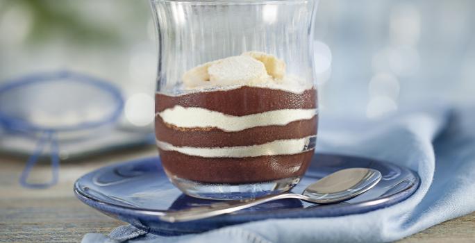 Fotografia em tons de azul e cinza, em uma bancada contém um pano azul e em cima um prato com um copo que contém açaí, banana e leite em pó. Ao lado uma colher para servir.