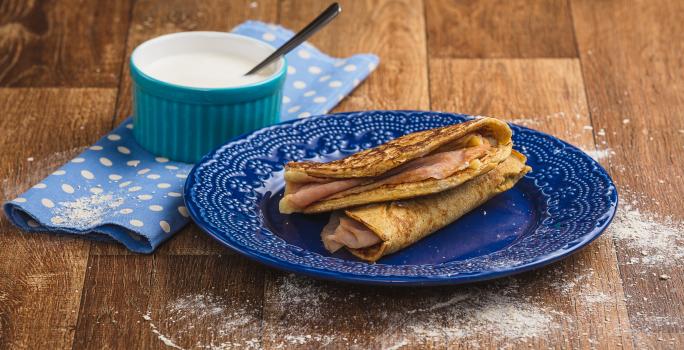 Imagem em tons de marrom e azul claro e escuro. No centro há um prato azul com duas quesadillas recheadas e, ao lado esquerdo, há um tecido azul com bolinhas brancas e, sobre ele, um pote azul com o creme azedo e uma colher.