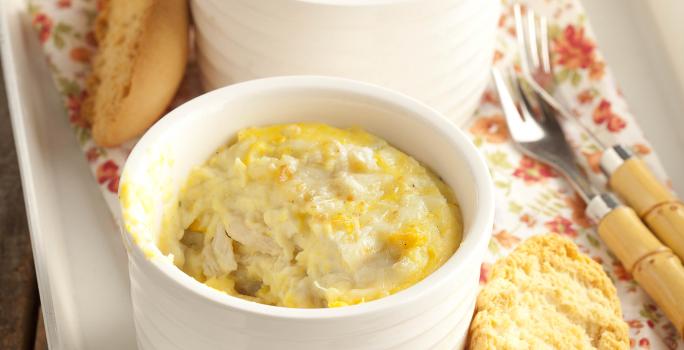 Fotografia em tons de amarelo em uma bancada de madeira escura, uma bandeja branca com um pano florido, duas cumbucas brancas com frango gratinado.