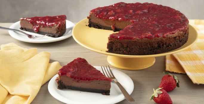 Fotografia em tons de amarelo em uma bancada de madeira com um suporte para bolo amarelo ao centro e a torta cheesecake de chocolate em cima. Ao lado, um paninho xadrez amarelo e dois pratos brancos com duas fatias da cheesecake de chocolate em cada um.