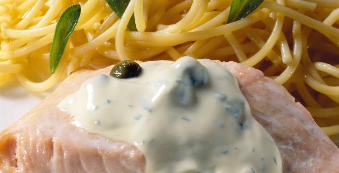 Uma mesa azul com bolinhas brancas vista de cima, um prato quadrado branco com macarrão espaguete, um pedaço de salmão com molho branco por cima.
