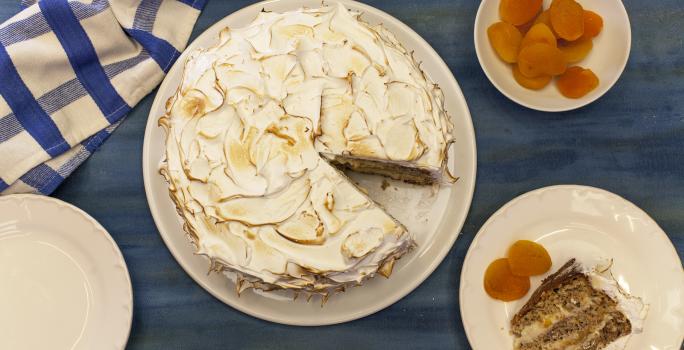 Fotografia em tons de azul e laranja em uma bancada azul, com um pano xadrez azul, um prato branco redondo com a torta de nozes em cima dele. Ao lado, um prato menor com a fatia da torta e damascos ao lado dele.
