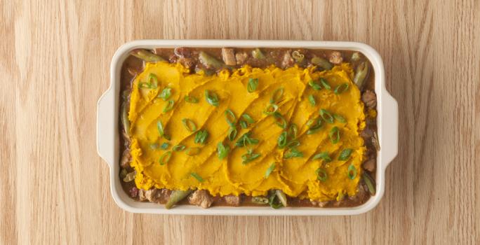 fotografia em tons de marrom e amarelo de uma bancada marrom vista de cima, contém um recipiente branco com alças com o pedaços de carne, purê de abóbora e cebolinhas por cima