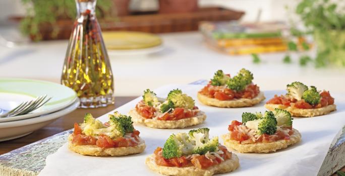 Fotografia em tons de amarelo e verde em uma bancada de madeira, com uma mesa arrumada para um jantar. Ao centro, uma tábua de madeira com um papel manteiga e várias mini pizzas com tomate e brócolis em cima dele.