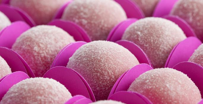 Fotografia em tons de rosa, com docinhos rosa claro na forma de bolinhas, envoltos em açúcar e dentro de forminhas de papel cartão na cor pink.