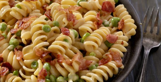 fotografia em tons de preto e bege de uma bancada preta vista de cima. Contém um recipiente preto redondo com macarrão parafuso, pedaços de bacon e ervilhas. fotografia em tons de preto e bege de uma bancada preta vista de cima. Contém um recipiente preto redondo com macarrão parafuso, pedaços de bacon e ervilhas.