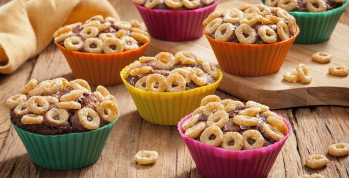 Fotografia em tons de laranja, verde, amarelo e rosa em uma bancada de madeira clara, um paninho amarelo, uma tábua de madeira, e várias forminhas de doces coloridas com o cupcake de chocolate cobertos com cheerios.