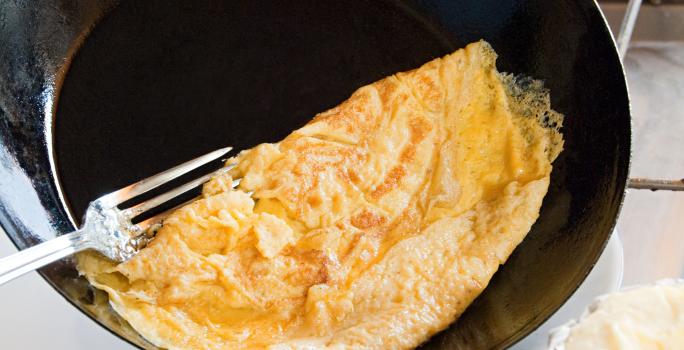 Fotografia de uma omelete dobrada em uma frigideira funda preta que está inclinada para colocar o ovo no prato, e um garfo segundo a omelete. Ao fundo, ovos sobre uma mesa branca. Ao lado do prato que vai receber a omelete, um pano de mesa vermelho.