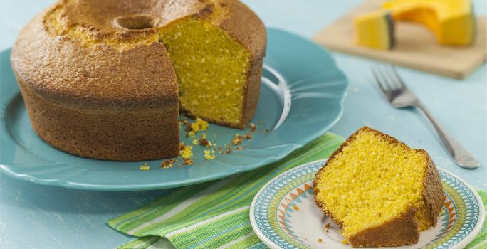 Fotografia em tons de azul de uma bancada azul com um prato azul, sobre ele um bolo de abóbora. Ao lado um paninho verde listrado com um prato redondo pequeno com uma fatia do bolo. Ao fundo uma tábua com uma fatia de abóbora.