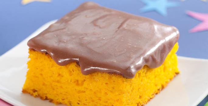 Fotografia em tons de azul, laranja e marrom de uma bancada azul, sobre ela um prato branco quadrado com uma fatia de bolo de cenoura com cobertura de chocolate. Ao lado um garfo verde. Ao fundo uma estrela azul e uma rosa.